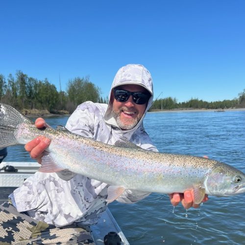 Smiling man in a hooded sun shirt and sunglasses holding a large rainbow trout over the side of a boat on the Kenai River under a clear blue sky.